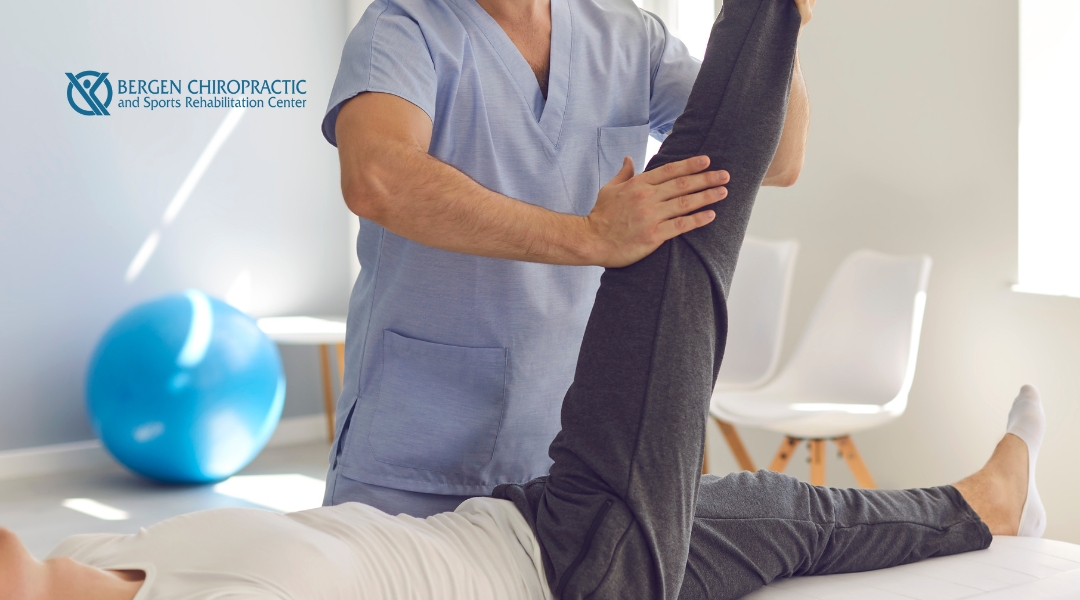 A physical therapist or chiropractor is assisting a patient lying on their back with a leg raised for a stretching exercise in a clinic setting.