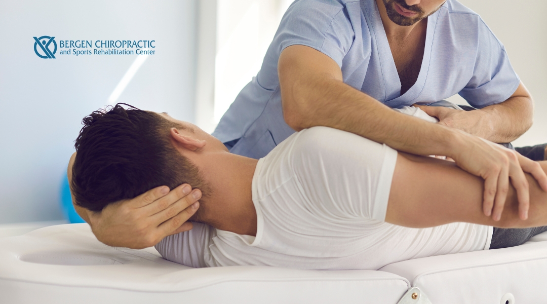 A chiropractor or manual therapist in blue scrubs is adjusting a patient's upper back and neck while the patient lies on their side.