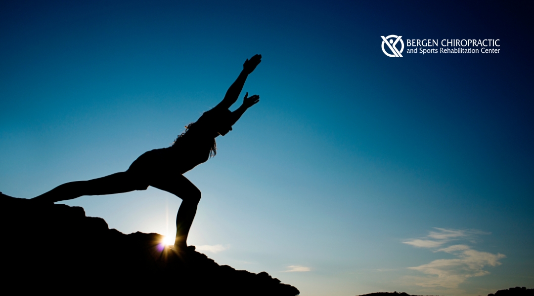 A silhouetted figure stands on a rocky peak with arms outstretched against a backdrop of mountains and a soft, hazy sky