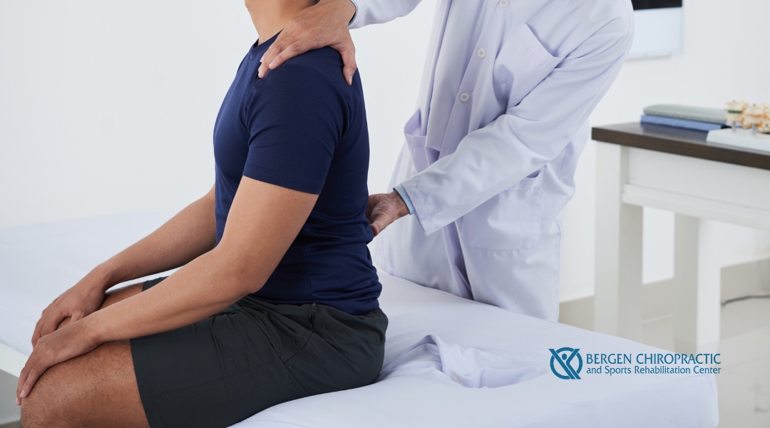 A chiropractor wearing a white coat gently adjusts the upper back of a seated patient dressed in a navy blue shirt and black shorts.