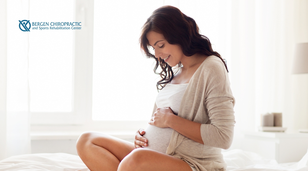 Smiling pregnant woman sitting on a bed in a peaceful, sunlit room with both hands gently resting on her baby bump.