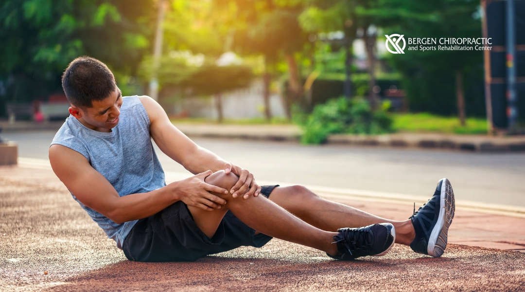 A man holding his knee while sitting on the ground after a possible sports injury, with a distressed look on his face. The background is a park with trees and a calm environment. The Bergen Chiropractic logo is visible in the corner.