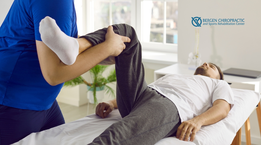 A chiropractor performing a leg stretch on a patient, assisting with rehabilitation in a bright clinic room. The patient is lying on a treatment table, and the chiropractor's hands are gently holding the leg. The Bergen Chiropractic logo is visible in the corner.