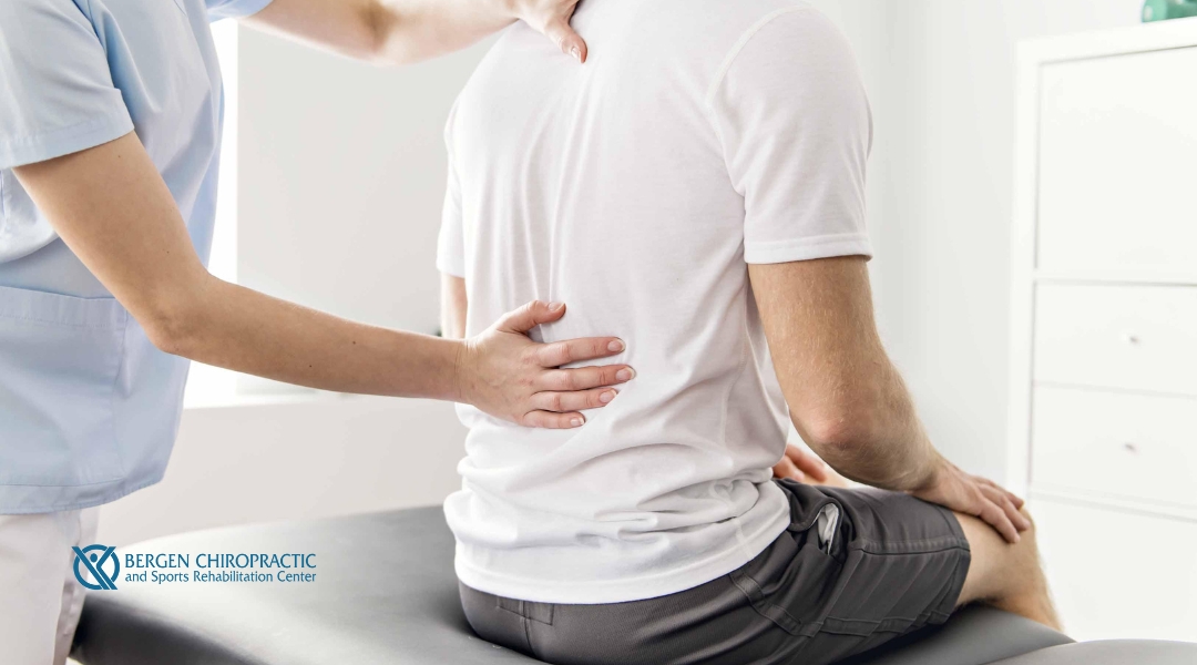 A chiropractor adjusting a patient's back while the patient sits on a treatment table. The chiropractor’s hand rests on the patient’s upper back, providing support during an adjustment. The Bergen Chiropractic logo is visible in the corner.