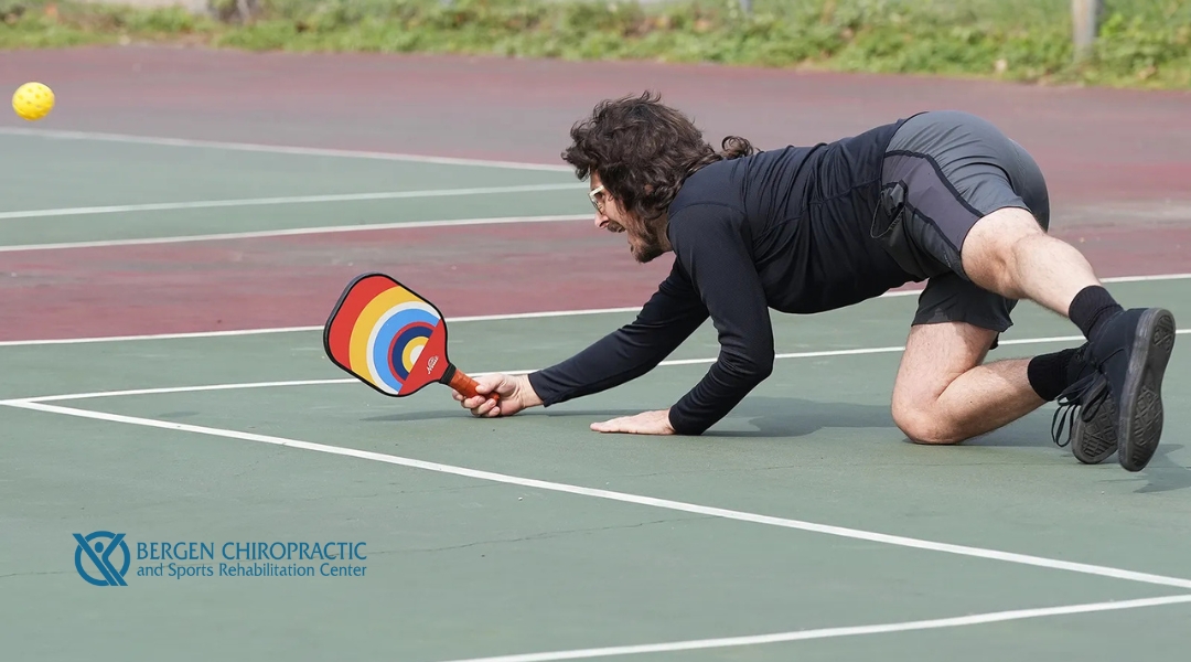 A man in black athletic wear dives low to the ground to hit a pickleball with a rainbow-colored paddle. He is on one knee, extending his arm fully. The Bergen Chiropractic and Sports Rehabilitation Center logo appears in the corner.