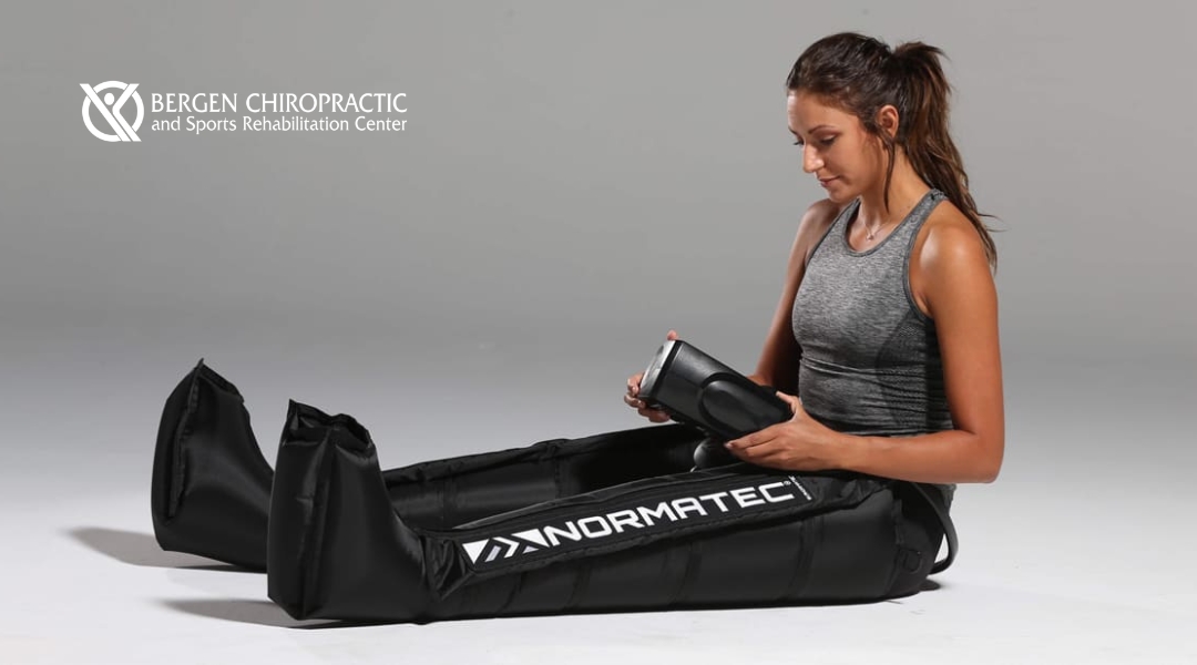 A female athlete sits on the floor wearing Normatec leg recovery boots and holding the control unit, ready to start compression therapy at Bergen Chiropractic.