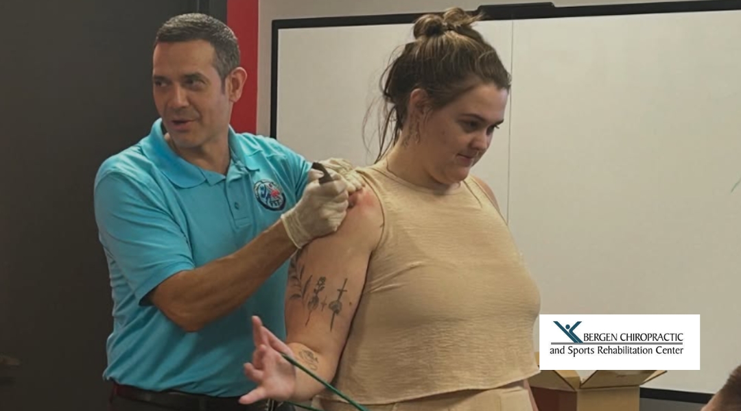A chiropractor demonstrates a technique on a patient’s shoulder while a group of onlookers is present, observing the procedure.